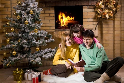 Portrait of cheerful family sitting on white fur reading old book with decorated fir tree near by