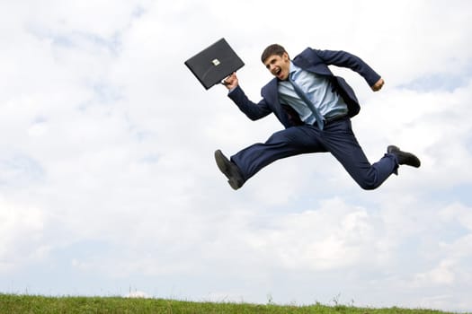 Handsome businessman in suit leaping over green grass with cloudy sky at background
