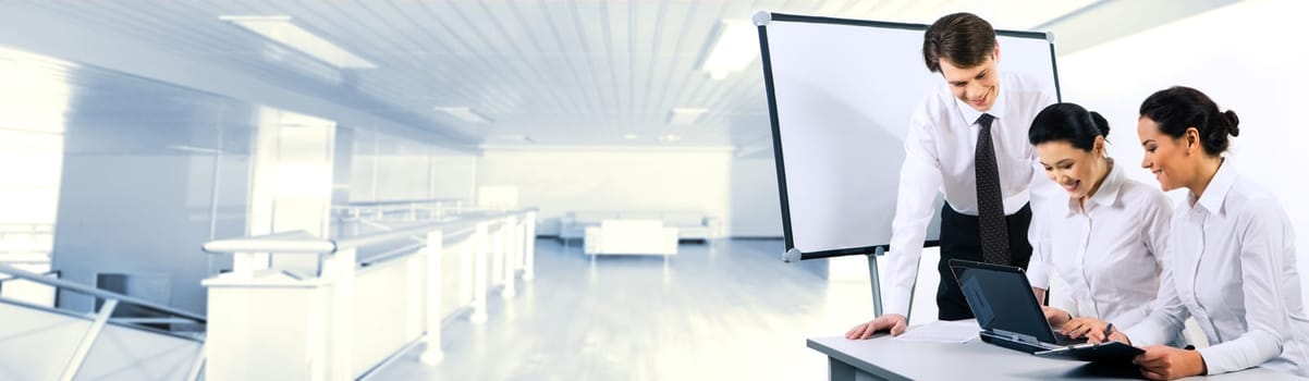 Two smiling business ladies sitting at table and looking at laptop monitor while their partner standing near by