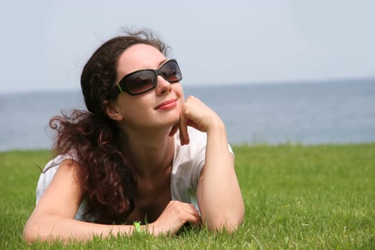 Portrait of pretty young lady relaxing on green grassland and looking aside through sunglasses during vacation