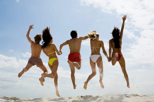 Rear view of friends holding by hands and jumping on sandy beach against sky