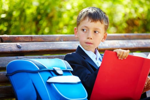 Portrait of smart boy sitting in the park with interesting book and looking aside