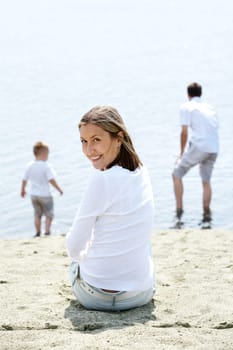 Image of smiling female sitting on sandy shore and looking at camera