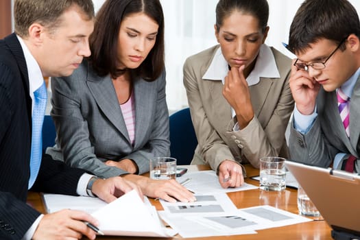 Photo of business group sitting at the table and watching at documents on it with pensive expression
