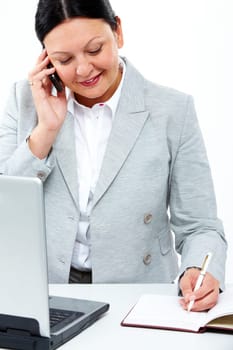 Busy secretary speaking on the phone and making notes in notepad with laptop near by