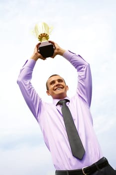 Portrait of happy businessman raising championship cup with cloudy sky above him