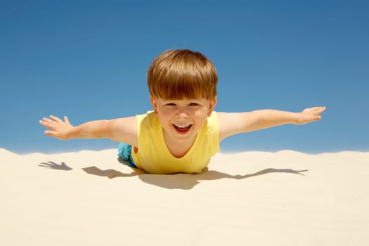 Happy boy lying on sand with outstretched arms and looking at camera on beach