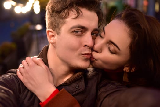 A young trendy couple in love walks down a night street and takes selfies. The girl is kissing her boyfriend.