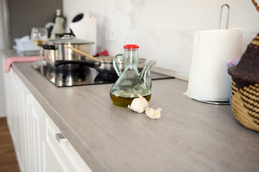 Food still life with a bottle of fresh extra virgin olive oil and garlic cloves on the table in the home kitchen countertop against saucepan on the electric stove. Cooking concept