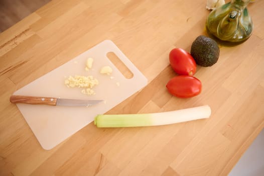 View from above of a chopped garlic clove on the cutting board, fresh organic vegetables for salad and oilcan with extra virgin olive oil on a wooden kitchen table with free advertising space