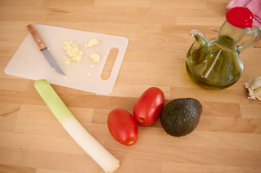 Chopped garlic on a cutting board, fresh organic leek, juicy red tomatoes and ripe avocado fruit near a bottle with olive oil on a wooden kitchen counter. Still life with healthy food. Copy ad space