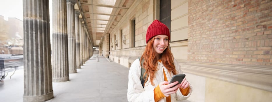 Smiling redhead girl in red hat, looks at map on smartphone app, checks her location, searches for a hostel. Tourist searches for popular attractions on mobile phone.
