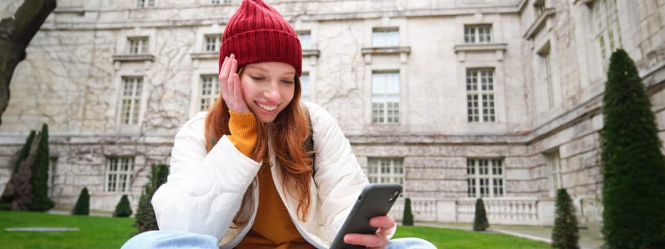 Portrait of stylish young woman, 25 years, sits on bench in park and uses mobile phone, reads online news, messages or watches video on smartphone app, connects to public wifi.