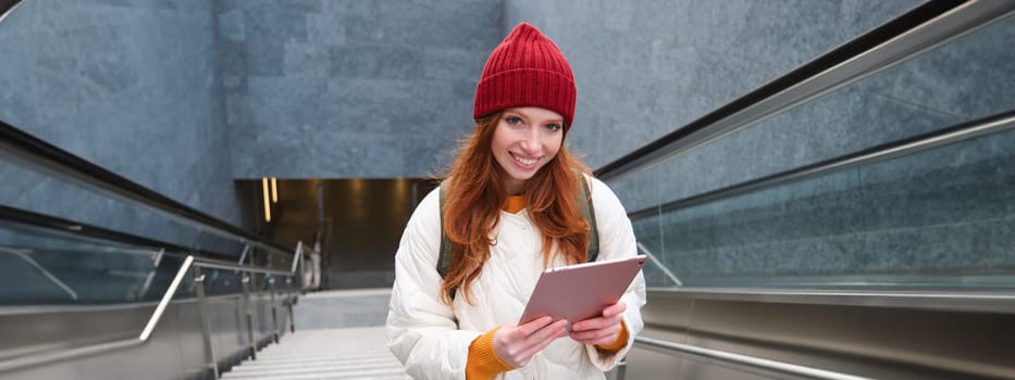 Smiling redhead woman walking up stairs with digital tablet, using gadget application while going somewhere in city.