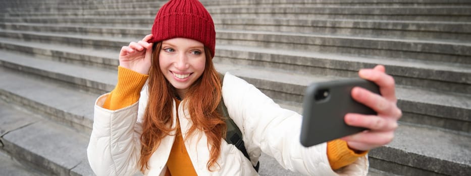 Stylish young girl in red hat, takes photos on smartphone camera, makes selfie as she sits on stairs near museum, posing for photo with app filter.