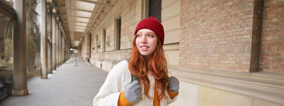 Backpack tourist. Happy redhead girl walks in city with a bag, goes on trip, explores city, wears red hat and gloves.