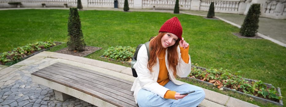 Young smiling redhead girl sits on bench and uses smartphone app, reads news online, watches video on mobile phone while relaxed in park.