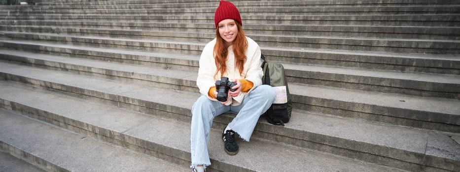 Young student, photographer sits on street stairs and checks her shots on professional camera, taking photos outdoors.