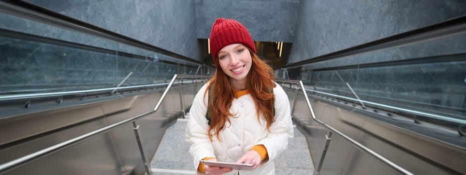 Beautiful redhead female model posing in city, walking up stairs with digital tablet, using gadget to plan her route, reading while going somewere.