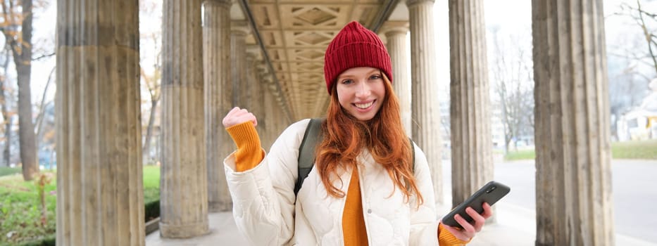 Smiling redhead girl looks excited, tourist holding mobile phone and does fist pump, thrilled about her journey, using smartphone on street.