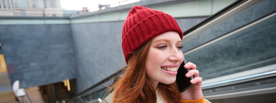 Portrait of happy redhead woman walking around town with smartphone, calling someone, talking on mobile phone outdoors.