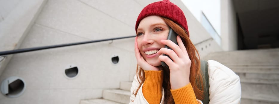 Beautiful smiling redhead female model, sits on street and talks on mobile phone, uses smartphone app to call abroad, laughing during telephone conversation.
