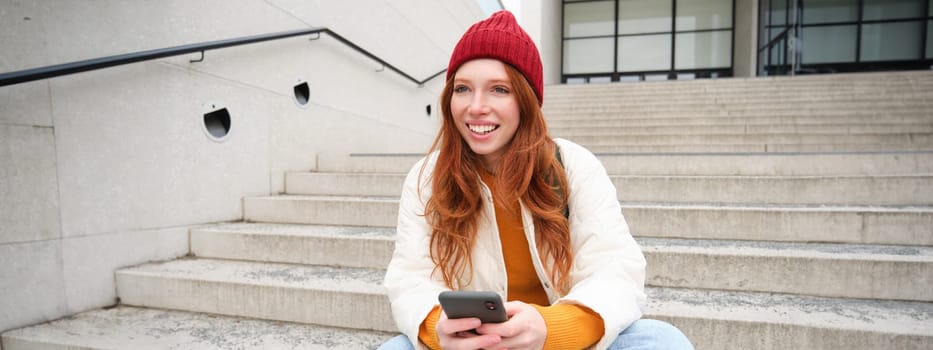 Redhead girl in red hat, sits on stairs and uses mobile phone. Modern woman holding smartphone, texting message, using telephone application outdoors.