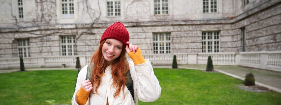 Beautiful redhead woman with backpack, tourist walking around city park and looks around, wears red hat and warm coat.