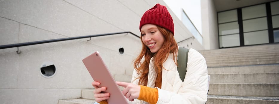 Happy stylish redhead girl, student in red hat, holds digital tablet, uses social media app, searches something online, connects to wifi.