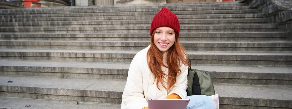 Beautiful young modern girl with red hair, holds digital tablet, sits on stairs near museum and connects public internet, sends message on gadget app.