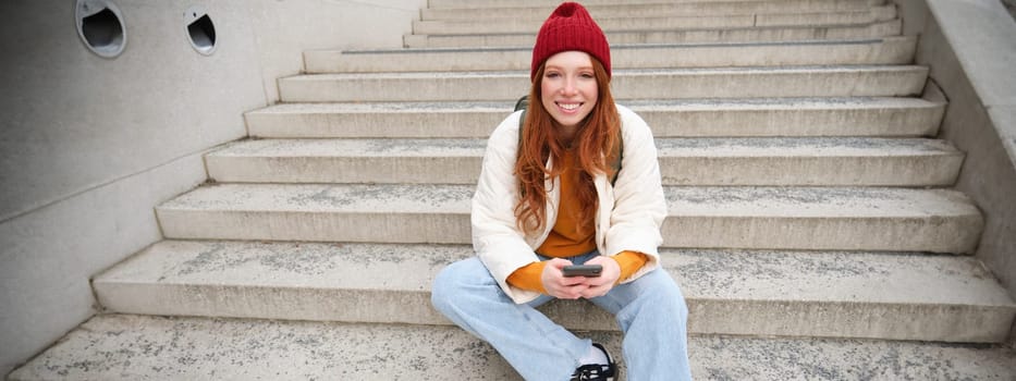 Hipster ginger girl, redhead woman sits on stairs with smartphone, waits for someone and messages on social media on mobile phone app. People and technology concept