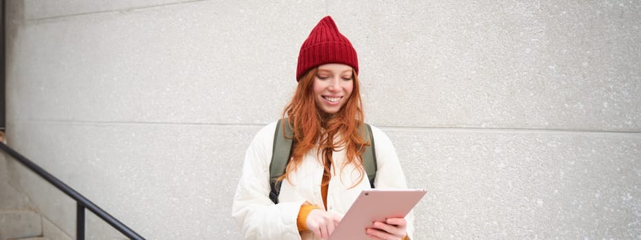 Young redhead woman with red hat, uses her digital tablet outdoors, stands on street with gadget, connects to wifi internet and searches for a location in internet.