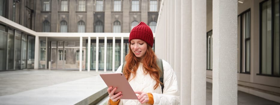 Happy redhead girl in red hat, walks around city with digital tablet, connects to public internet wifi and looks for route, looks at map on her gadget.