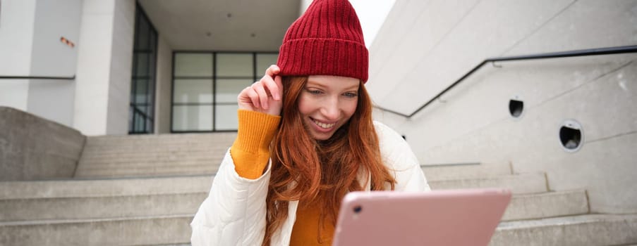 Beautiful redhead female model, ginger girl with digital tablet, sits on stairs outdoors, reads on her gadget, uses internet application and wifi on street.