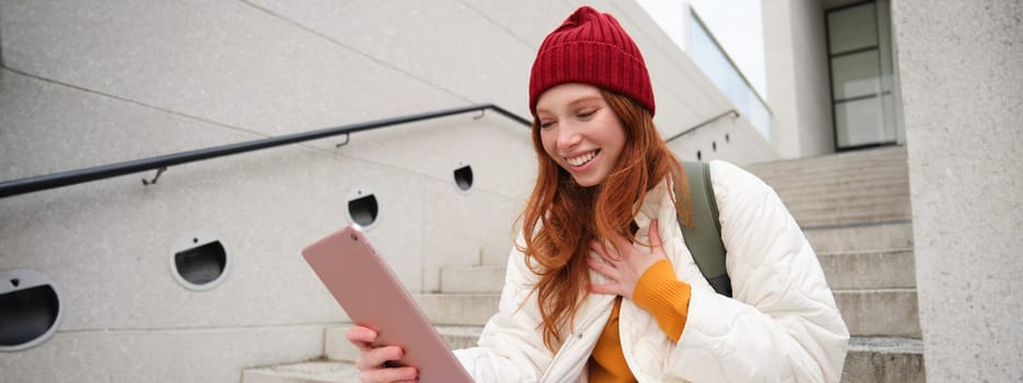 Happy stylish redhead girl, student in red hat, holds digital tablet, uses social media app, searches something online, connects to wifi.