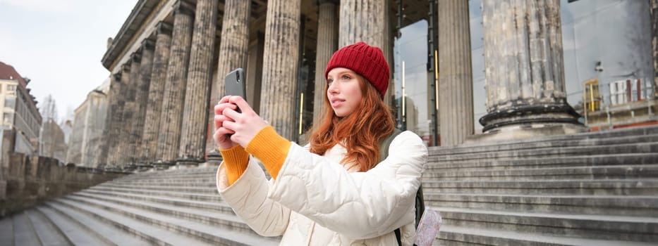 Portrait of young blogger, redhead girl tourist takes pictures of sightseeing, makes photos on smartphone camera, records video and smiles.