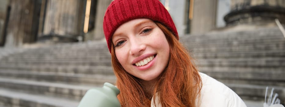 Young redhead female tourist rests during her trip, opens thermos and drinks hot tea, having a break after sightseeing.