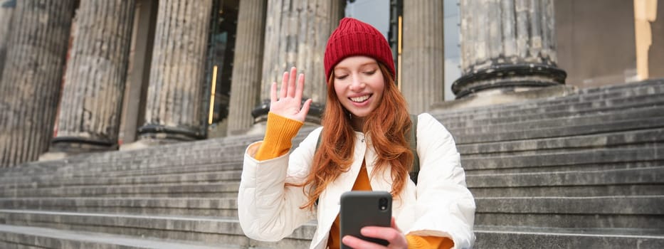 Young redhead woman sits on stairs outdoors and waves hand at smartphone camera, video chats with friends, connects to public wifi.