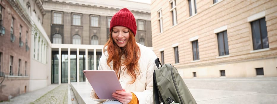 Redhead girl smiles, sits outdoors near building with digital tablet, thermos and backpack, connects to public internet and searches smth online on her gadget.