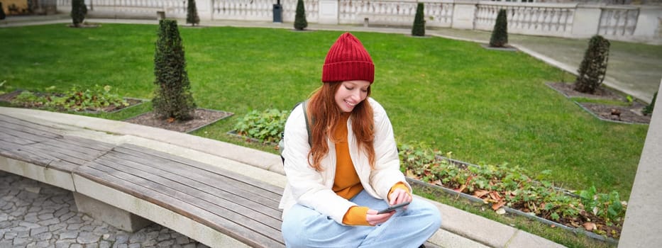 Portrait of stylish young woman, 25 years, sits on bench in park and uses mobile phone, reads online news, messages or watches video on smartphone app, connects to public wifi.