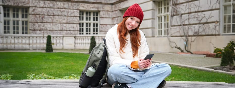 Redhead girl, female student sits with mobile phone on bench in parj, leans on her backpack. Woman browsing social media app feed on her smartphone.