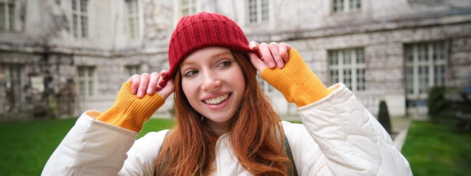 Portrait of smiling redhead woman puts on red hat and smiles, wearing warm clothes while exploring city streets.