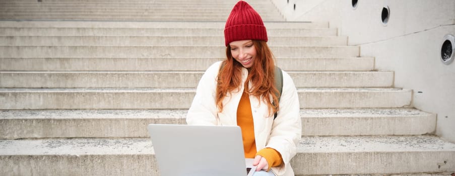 Smiling redhead girl, young woman typing on laptop keyboard, sitting outdoors on stairs with computer, working remote, doing her homework on fresh air.