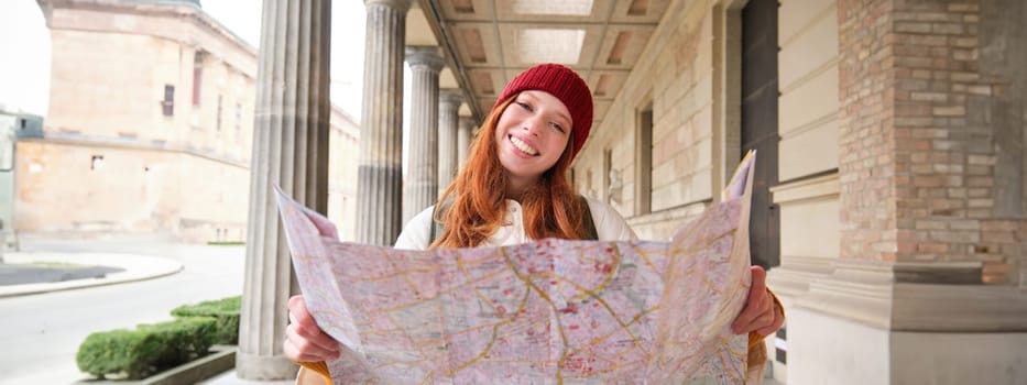 Adventurous redhead girl walks in town with paper map, explores city as tourist, looks for popular tourism attractions, looks around excited and smiles.