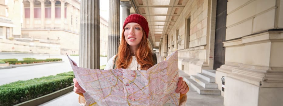 Adventurous redhead girl walks in town with paper map, explores city as tourist, looks for popular tourism attractions, looks around excited and smiles.