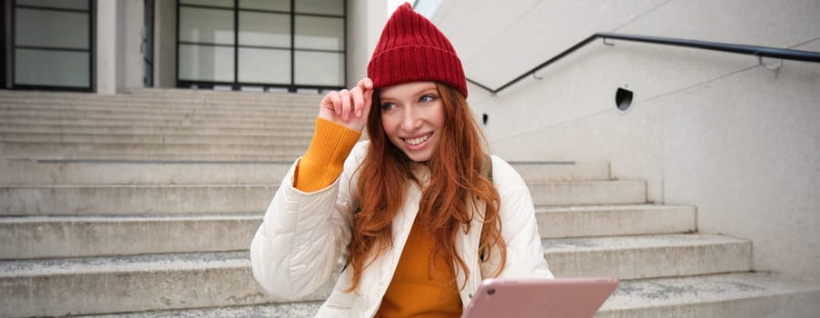 Beautiful redhead female model, ginger girl with digital tablet, sits on stairs outdoors, reads on her gadget, uses internet application and wifi on street.