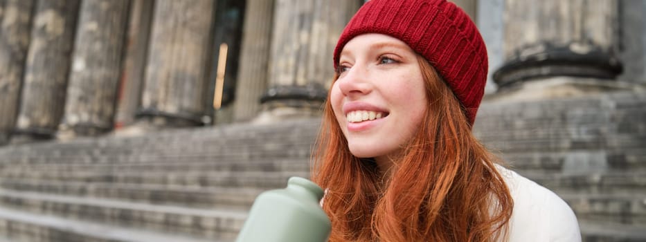 Smiling redhead girl in red hat, sits on stairs near building with flask, drinks water and looks happy.