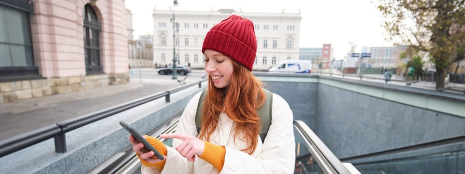 Portrait of young urban girl with red hair, using smartphone, texting message, using mobile phone application while going up an escalator.
