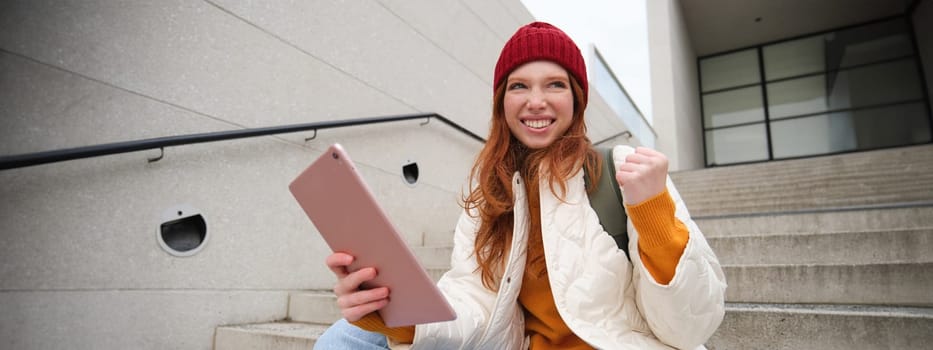 Happy stylish redhead girl, student in red hat, holds digital tablet, uses social media app, searches something online, connects to wifi.