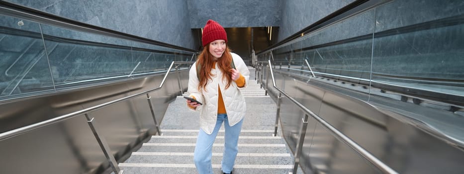 Outdoor shot of young woman plans route, follows map on smartphone app, goes up stairs with backpack and smiles.